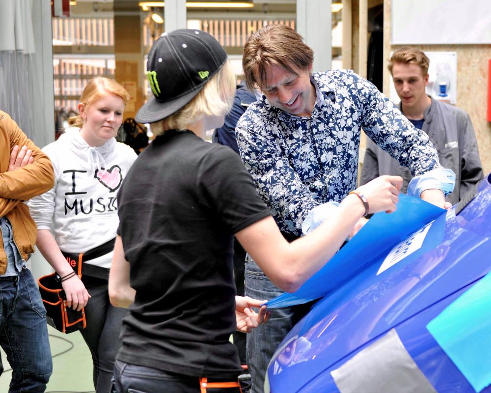 Trainer helpt student bij oefening folie wrappen op motorkap tijdens Appcademy workshop en gastcollege op ROC / MBO signopleiding