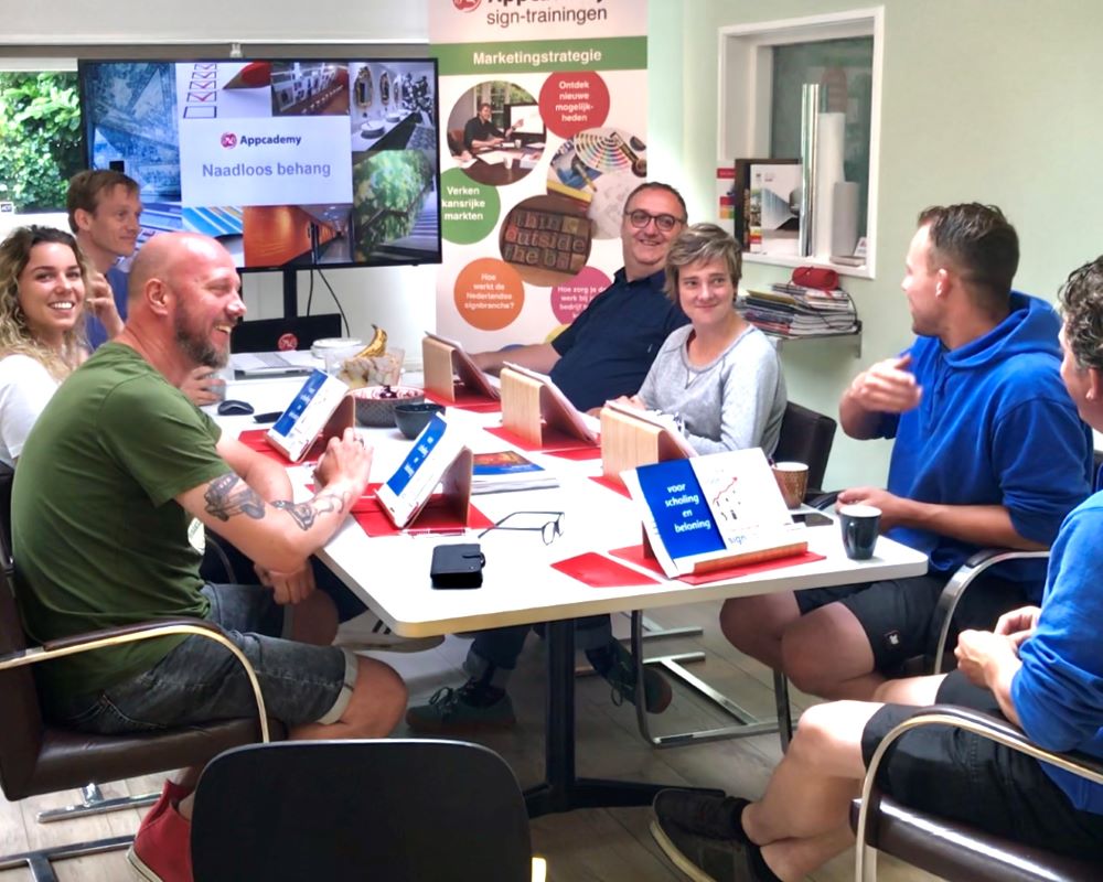 Gezelschap van trainer en cursisten praten gezellig aan tafel tijdens Appcademy Naadloos fotobehang workshop