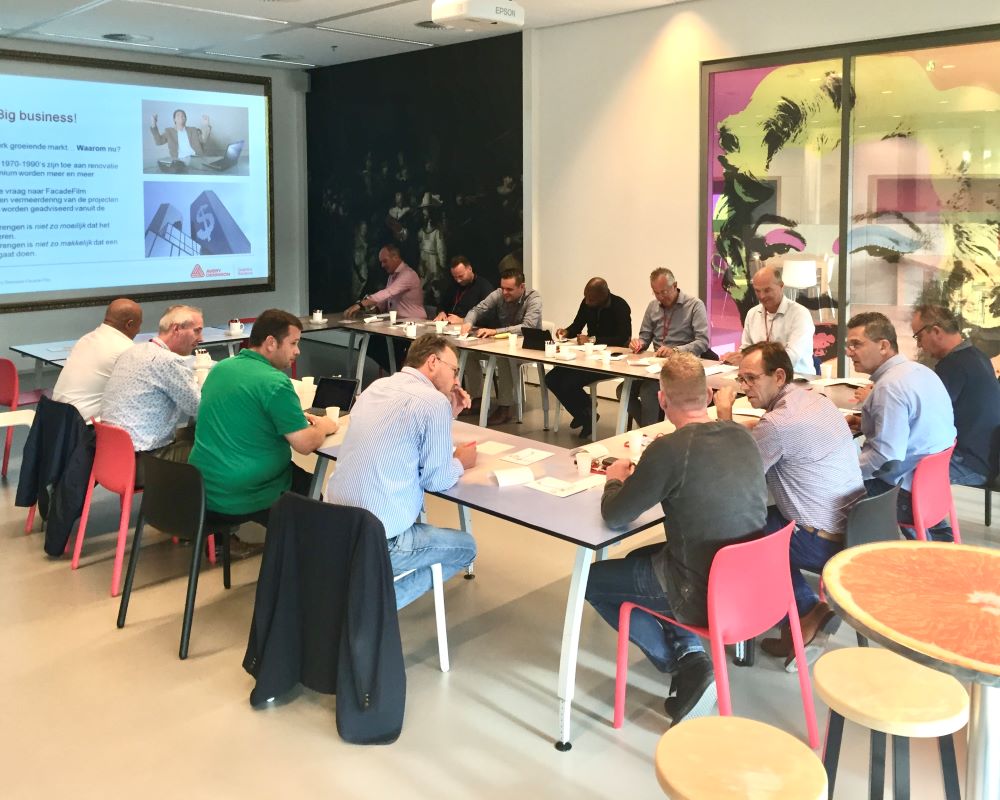 Gezelschap van trainer en grote groep cursisten aan tafel bij uitleg over kozijnfolie tijdens “Schilderen met folie” Facadefilm cursus en opleiding