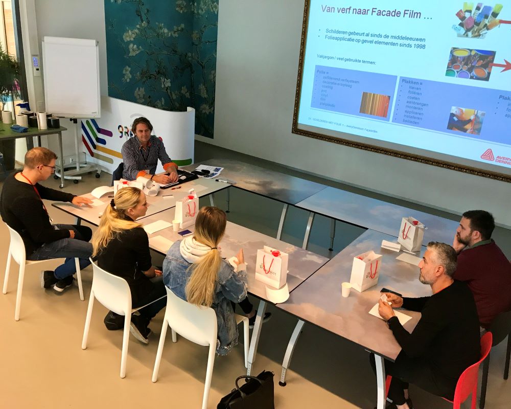 Gezelschap van trainer en 3 cursisten aan tafel bij theorie uitleg over kozijnfolie tijdens “Schilderen met folie” Facadefilm cursus